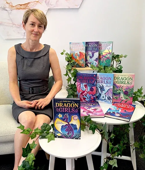 Meredith Badger, Teil des Autorinnen-Duos Maddy Mara, sitzt auf einem Sofa, neben sich auf zwei kleinen Tischen sind alle Dragon Girls-Bücher (Band 1-9) zu sehen